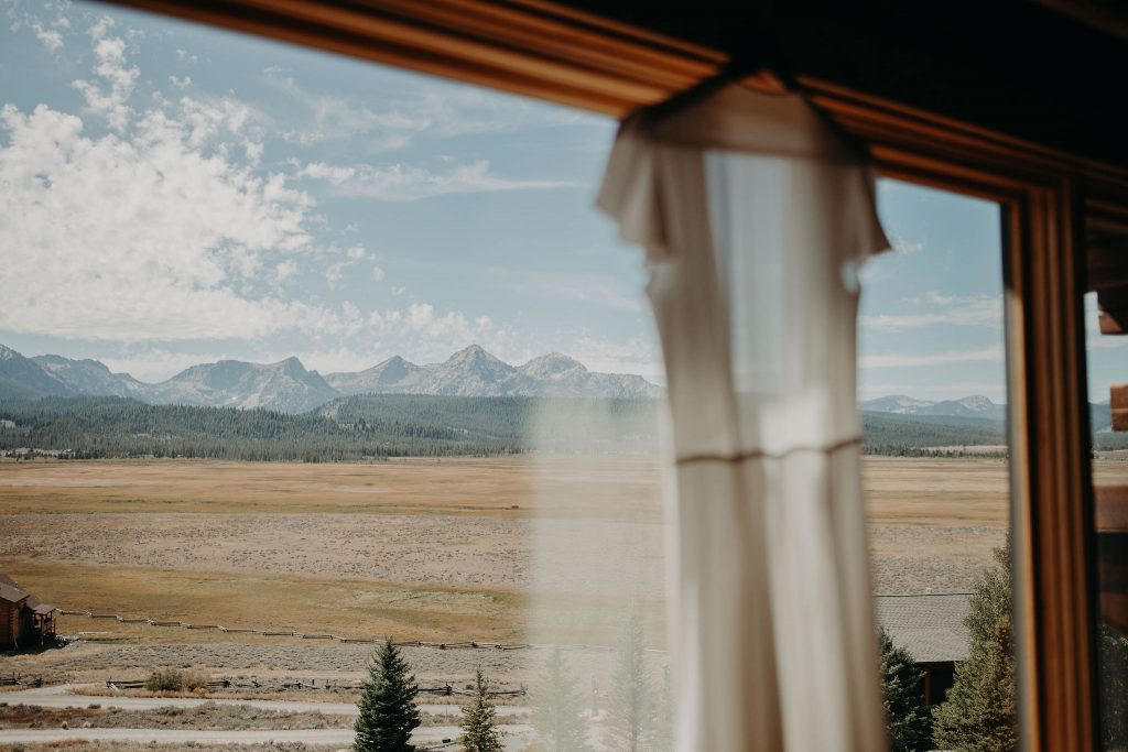 A brides wedding dress hanging on a window with views of the Sawtooth Mountains in the distance.