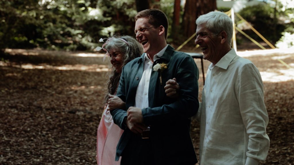 A groom walking down the wedding aisle with his parents under the redwoods at Old Mill Park.