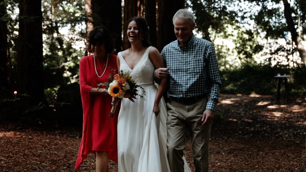 A bride walking down the wedding aisle with her parents under the redwoods at Old Mill Park.